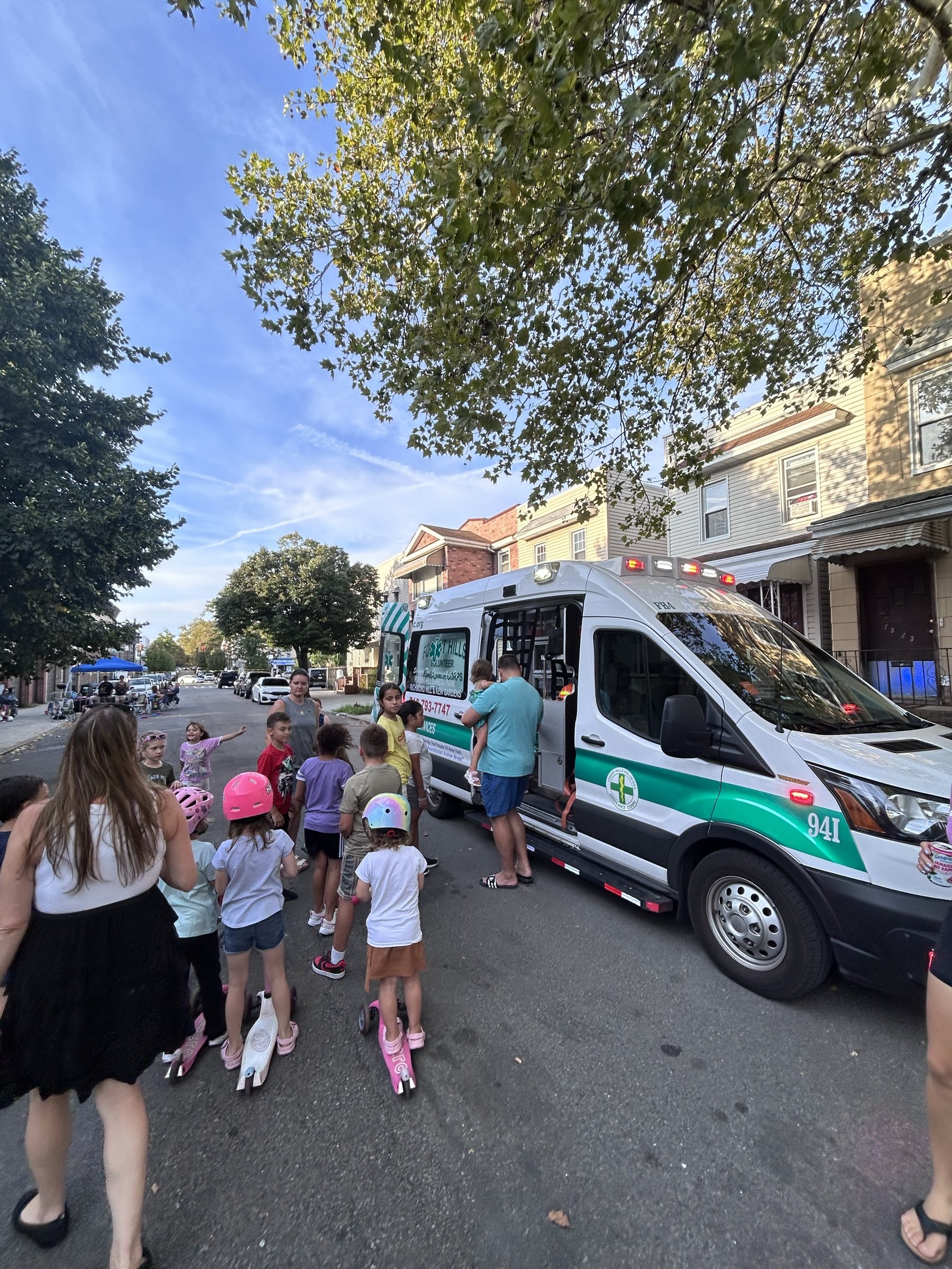 Children with scooters and bicycles crowded around FHVAC ambulance FH4 on a residential street during a block party, with tents set up further down the block